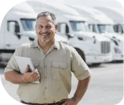 Homem mais velho com a mão na cintura olhando para a câmera e sorrindo, ele tem cabelo e barba meio grisalhos e ao fundo tem vários caminhões estacionados de uma transportadora